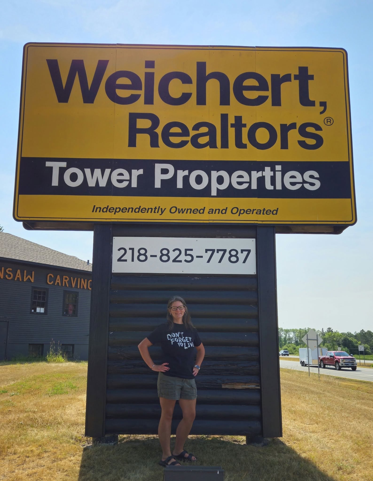 Person stands in front of a large “Weichert, Realtors Tower Properties” sign with a phone number. The person is smiling and wearing a dark shirt and shorts. The scene is outdoors on a sunny day.