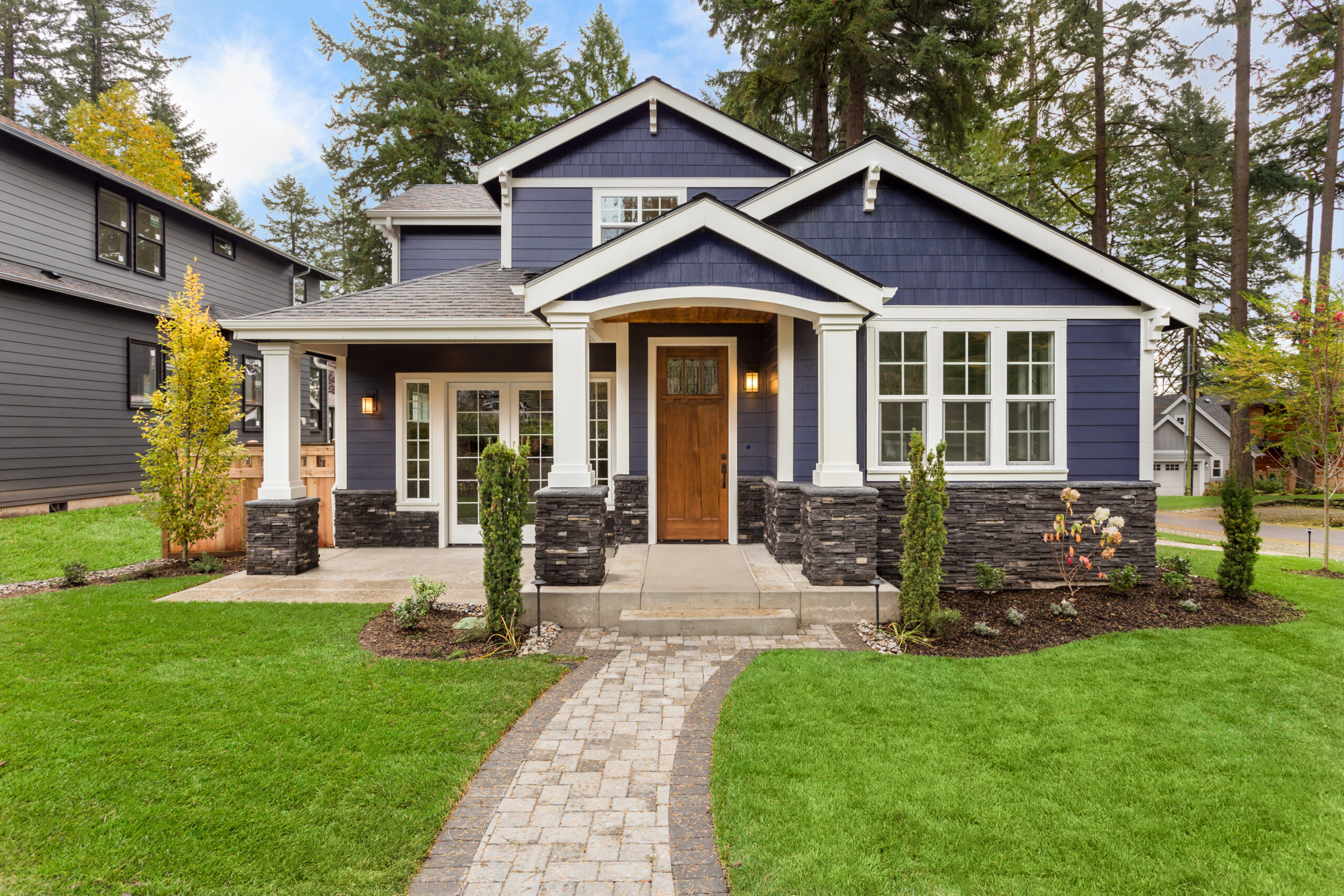 A modern blue house with white trim, stone accents, and a wooden front door, surrounded by a green lawn, small shrubs, and tall trees in the background. Brick pathway leads to the front porch.