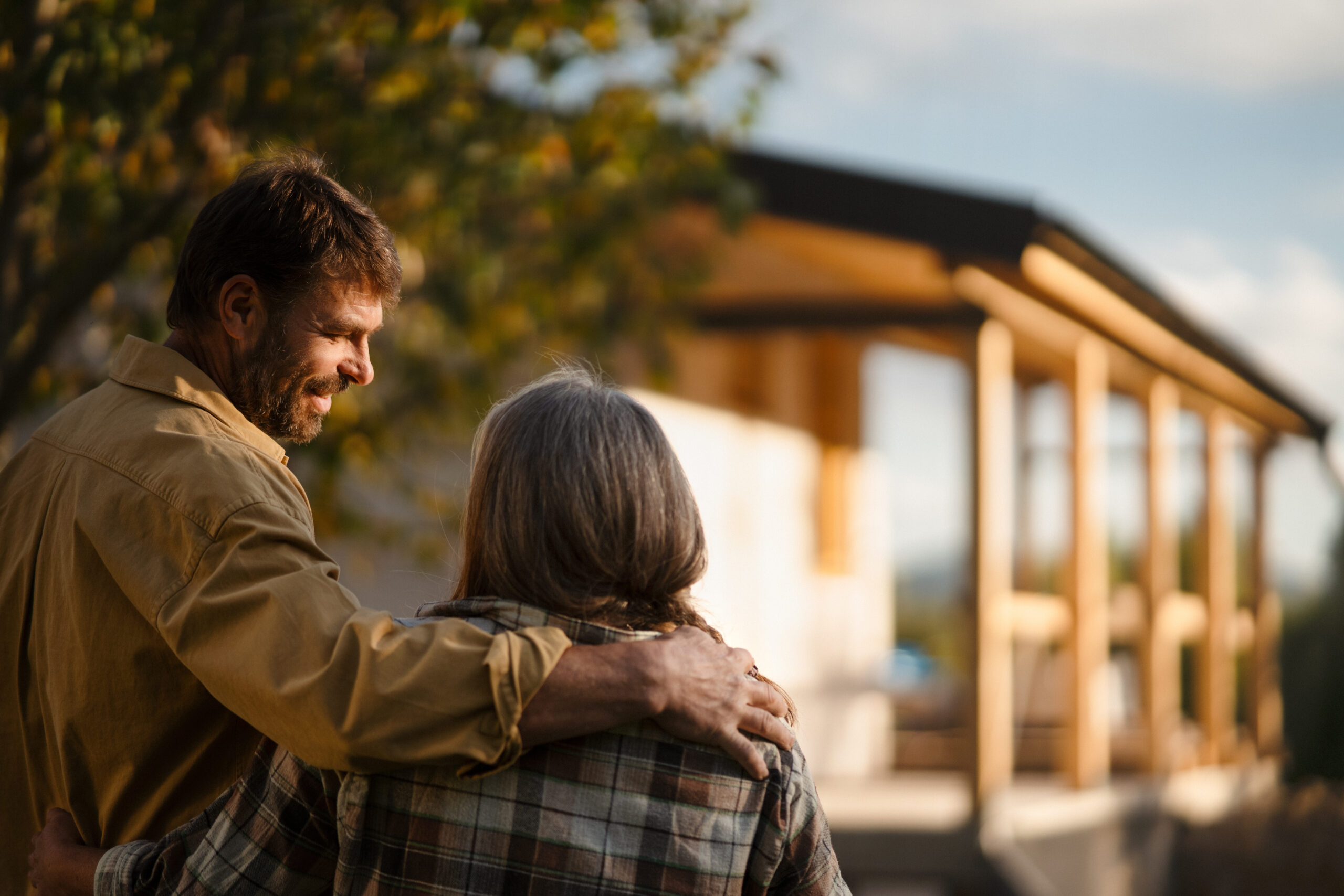 A man and an older woman stand outside, smiling, with his arm around her shoulders. A modern house and trees are in the background, bathed in warm sunlight.