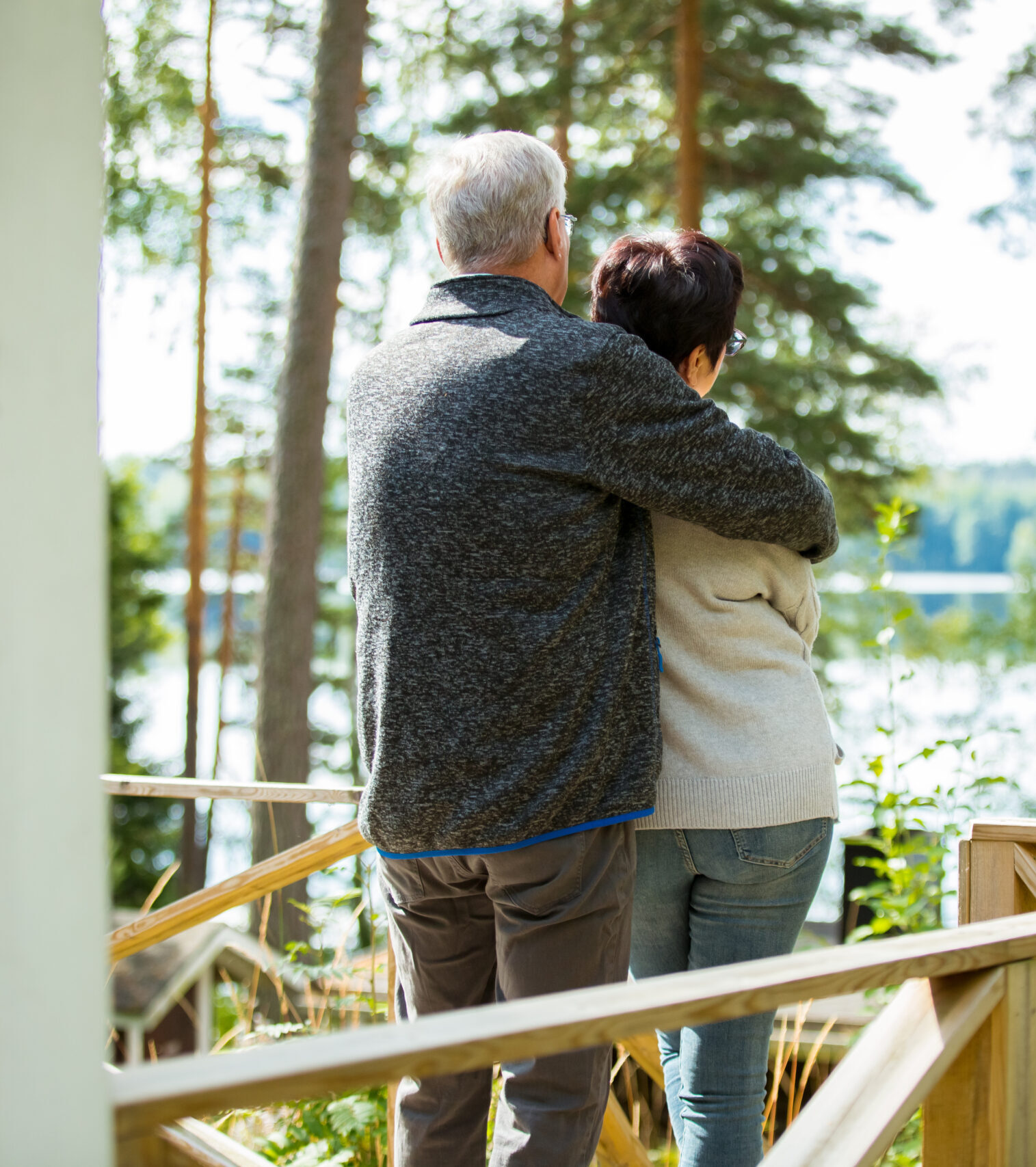 An older couple stands on a wooden porch, embracing and looking out at a lake surrounded by trees on a sunny day.