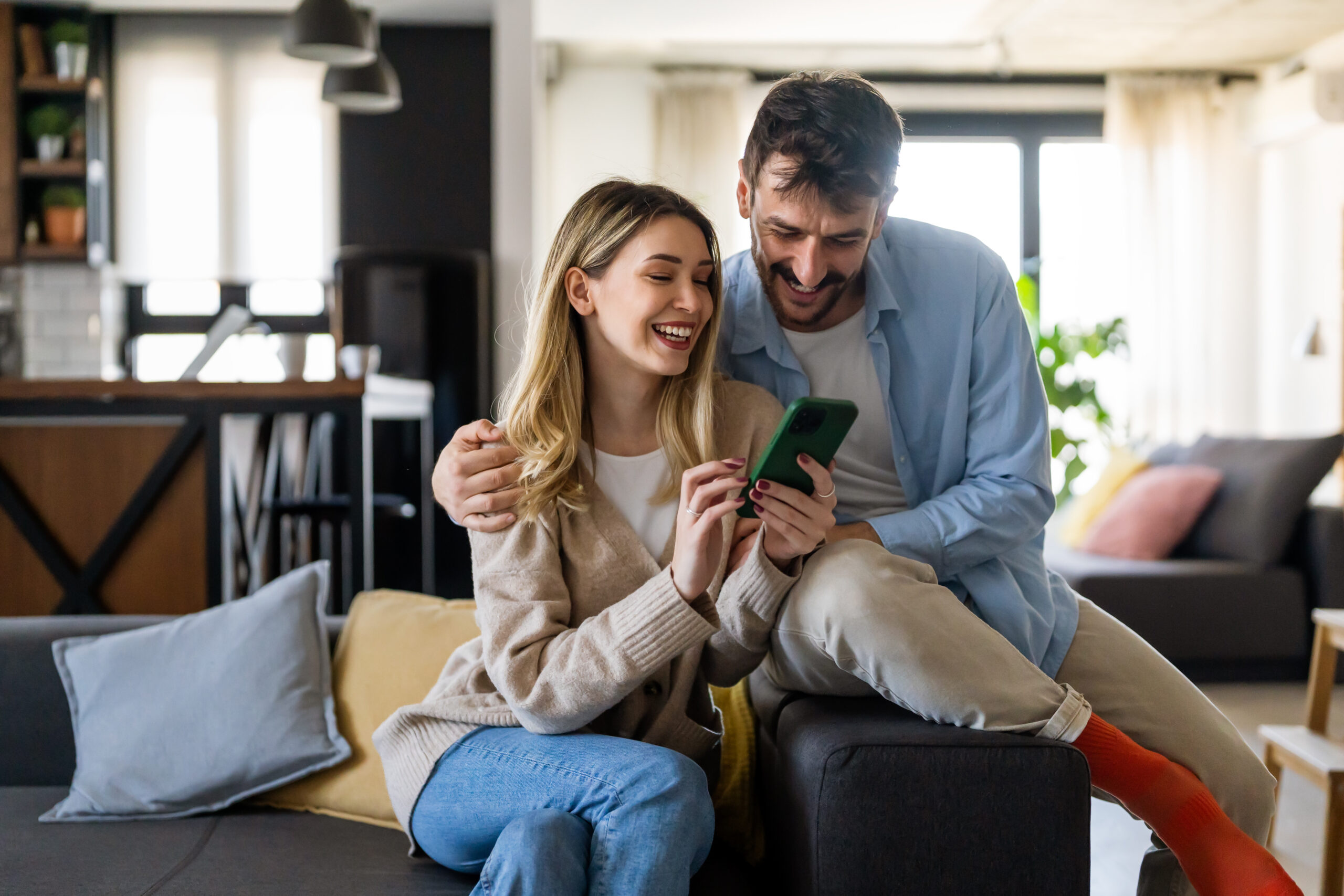 A smiling couple sits on a couch at home, looking at a smartphone together. The woman is holding the phone, and the man has his arm around her shoulders. The setting is cozy and well-lit.