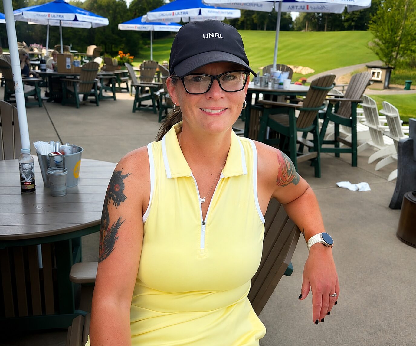 Amanda, a woman in a sleeveless yellow top and black cap, sits outdoors at a patio table, smiling. She has glasses, tattoos on both arms, and wears a smartwatch. Umbrellas and green grass are visible in the background.