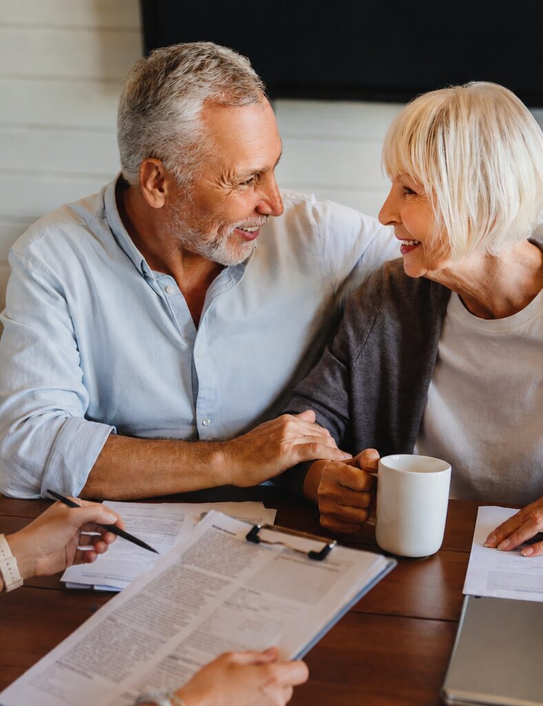 Smiling older couple sits at a table with documents, a clipboard, and a coffee mug, holding hands and looking at each other while another person’s hands are visible in the foreground—capturing a warm moment during the process of selling your home.