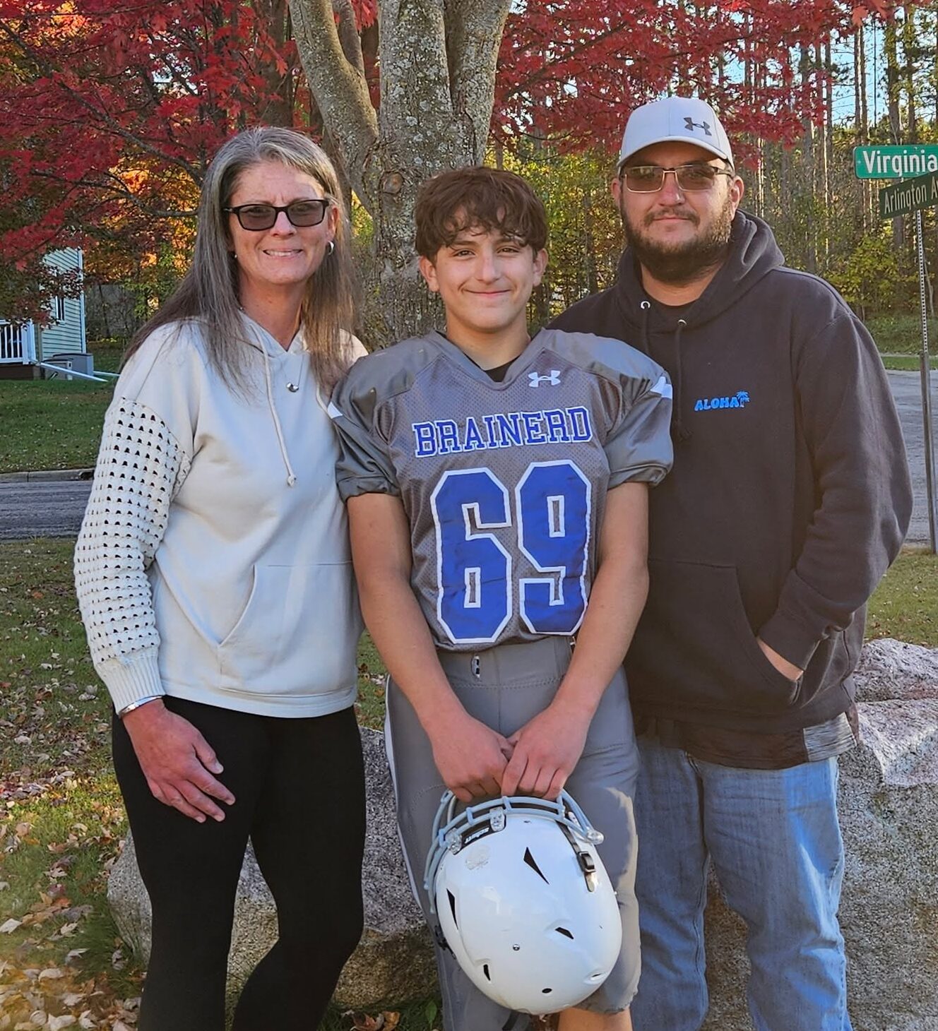 Three people stand outdoors: a woman in a hoodie, a boy in a football uniform holding a helmet, and a man in a hoodie and cap. Autumn trees and a green street sign are visible in the background.